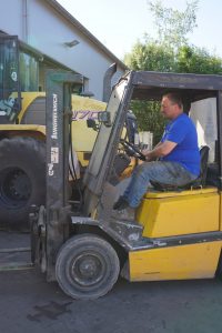 Mitarbeiter des Lohnbetriebs Bohmann fährt mit einem gelben Gabelstapler auf dem Hof vor einer großen Landmaschine.
