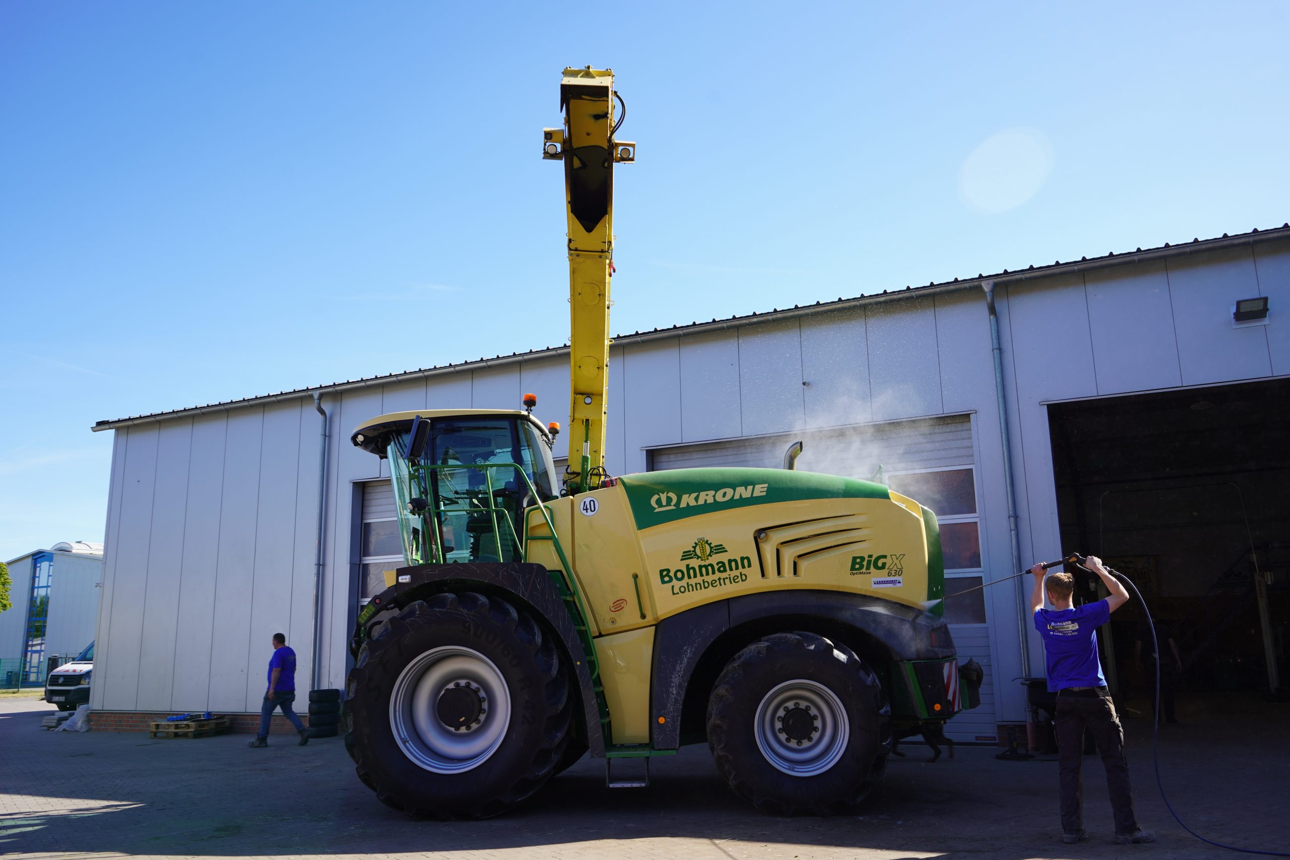 Mitarbeiter des Lohnbetriebs Bohmann reinigt mit Hochdruckreiniger einen Krone BiG X Feldhäcksler auf dem Hof.