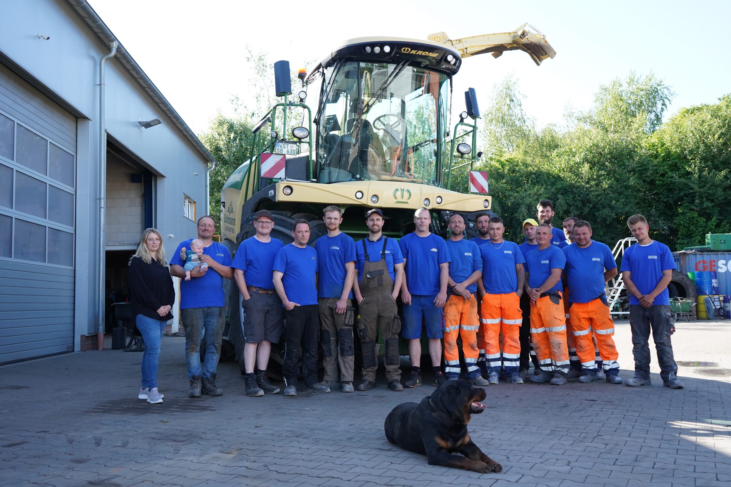 Gruppenfoto des Teams vom Lohnbetrieb Bohmann vor einem Krone-Feldhäcksler, mit Mitarbeitern, einer Frau mit Baby und einem Hund im Vordergrund.