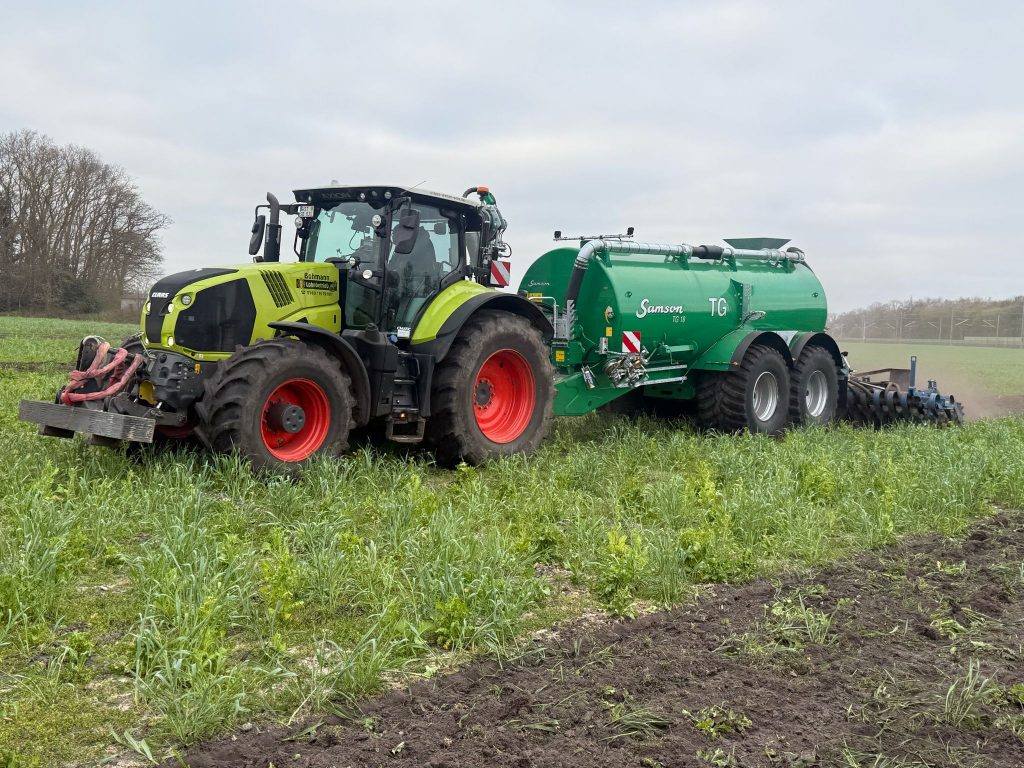 Traktor mit grünem Güllefass von Samson und Grubber im Einsatz auf einem Feld.