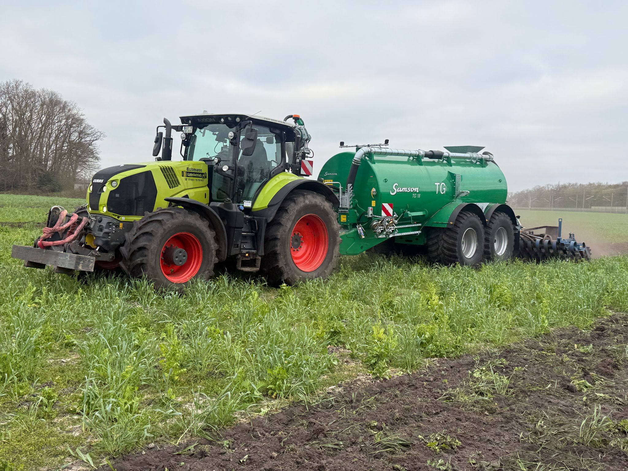 Traktor mit grünem Güllefass von Samson und Grubber im Einsatz auf einem Feld.
